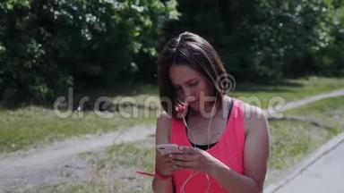 戴着耳机的可爱女孩穿过公园。 迷人的年轻女士戴着耳机在发短信。 看智能手机的女孩